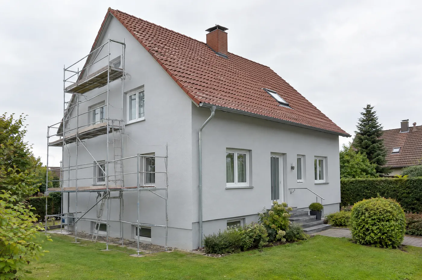 Einfamilienhaus mit frisch gestrichener, hellgrauer Fassade und rotem Ziegeldach, an einer Seite steht noch das Gerüst.